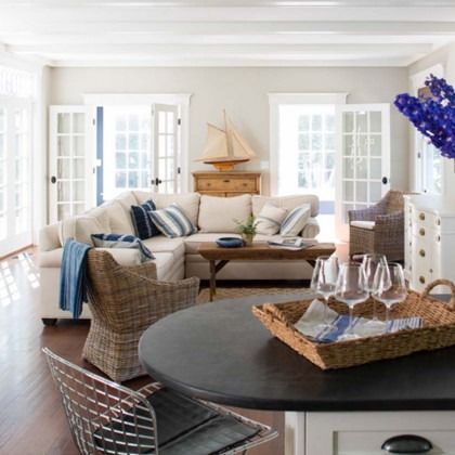 Bright coastal living room with white walls, beige sofa, wicker chairs, blue accents, and a dining table in front.
