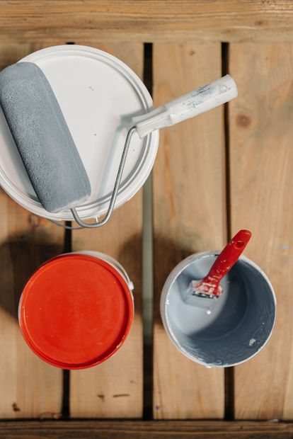 Paint roller, tray, and two paint cans on wooden floorboards with gray, red, and orange paint