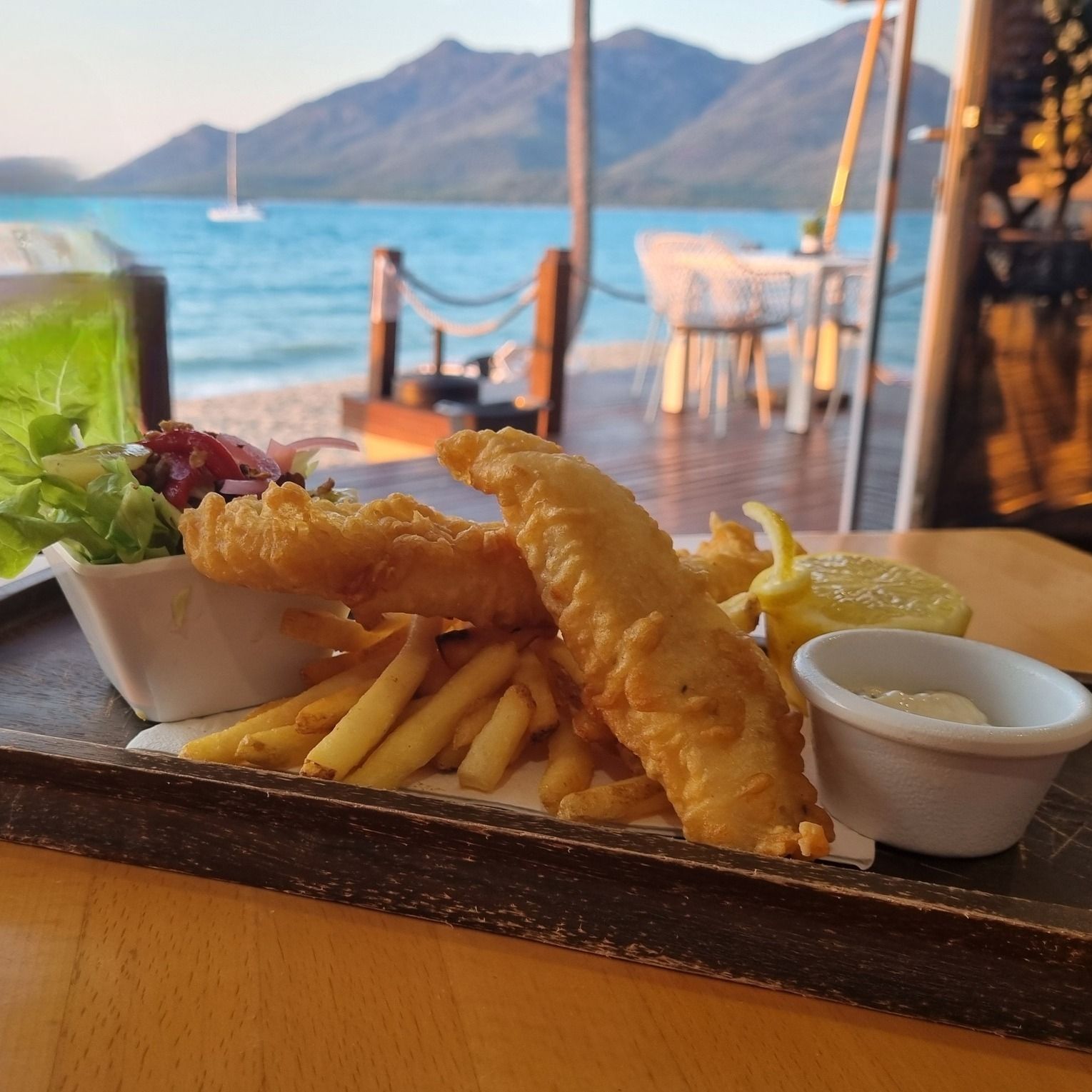 Fish and chips with salad and sauce, served outdoors with a beach and mountains in the background.