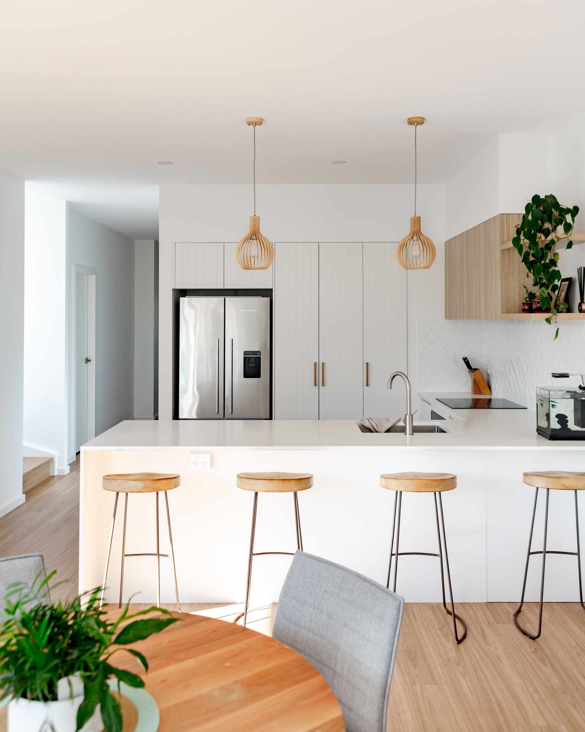Modern kitchen with white cabinets, wooden stools, and light fixtures; a round table with a chair and a plant.