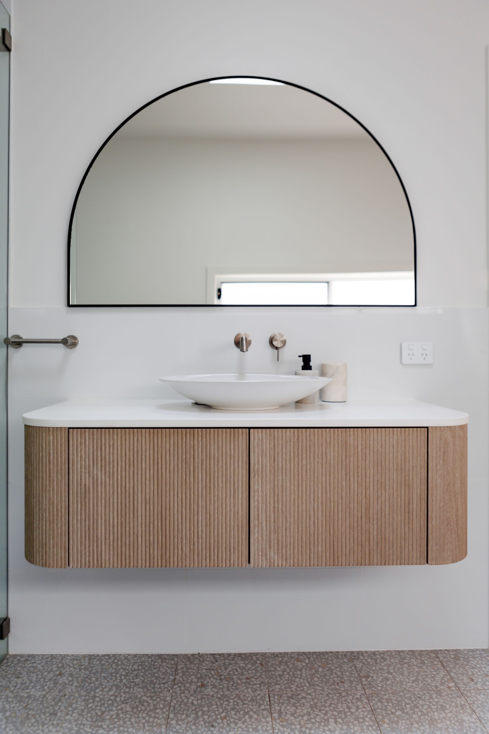 Floating wooden bathroom vanity with an arched mirror, white countertop, and vessel sink.