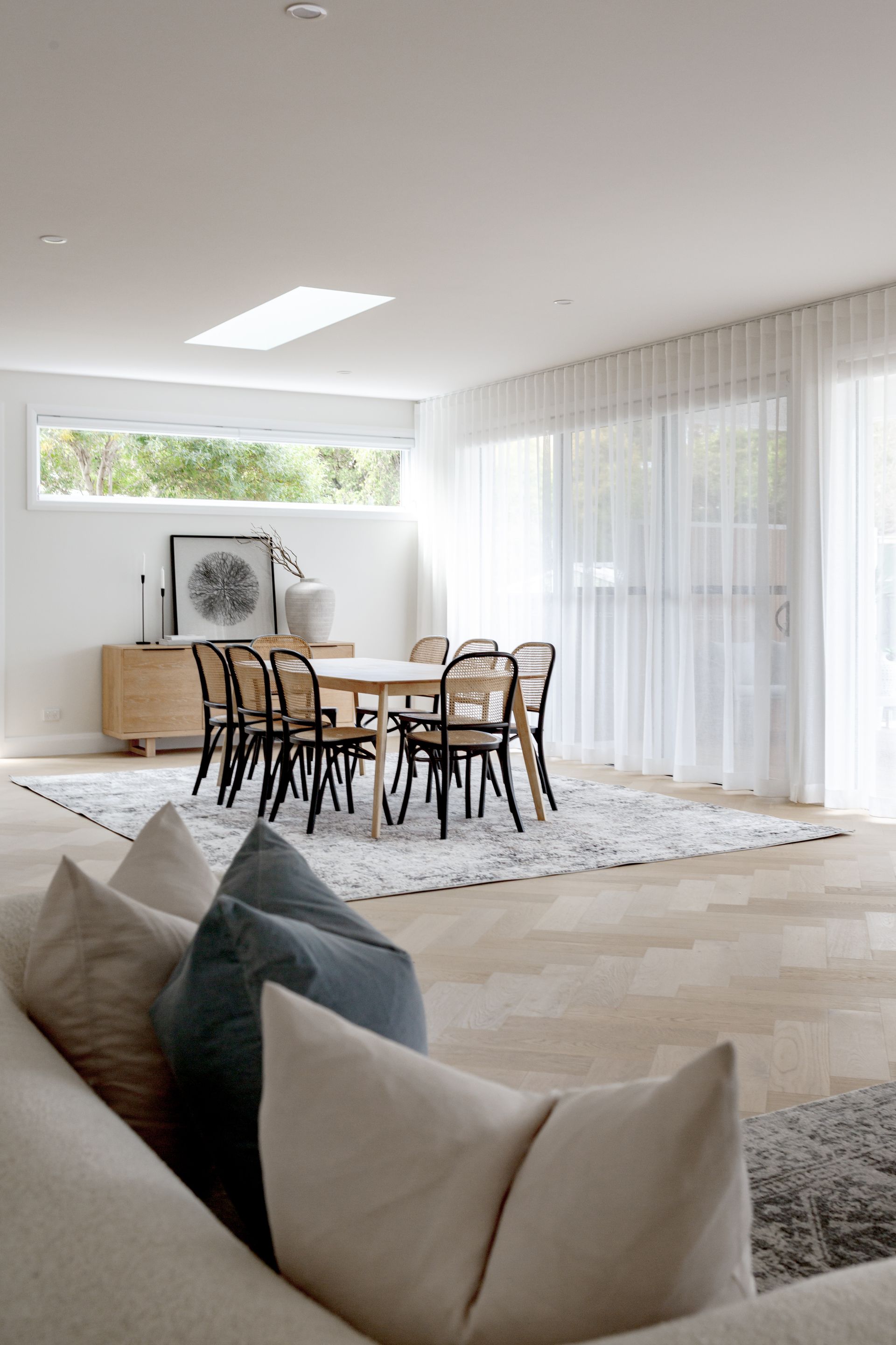 Open-plan dining area with a round table, chairs, and a rug, seen from a cream-colored sofa with pillows.
