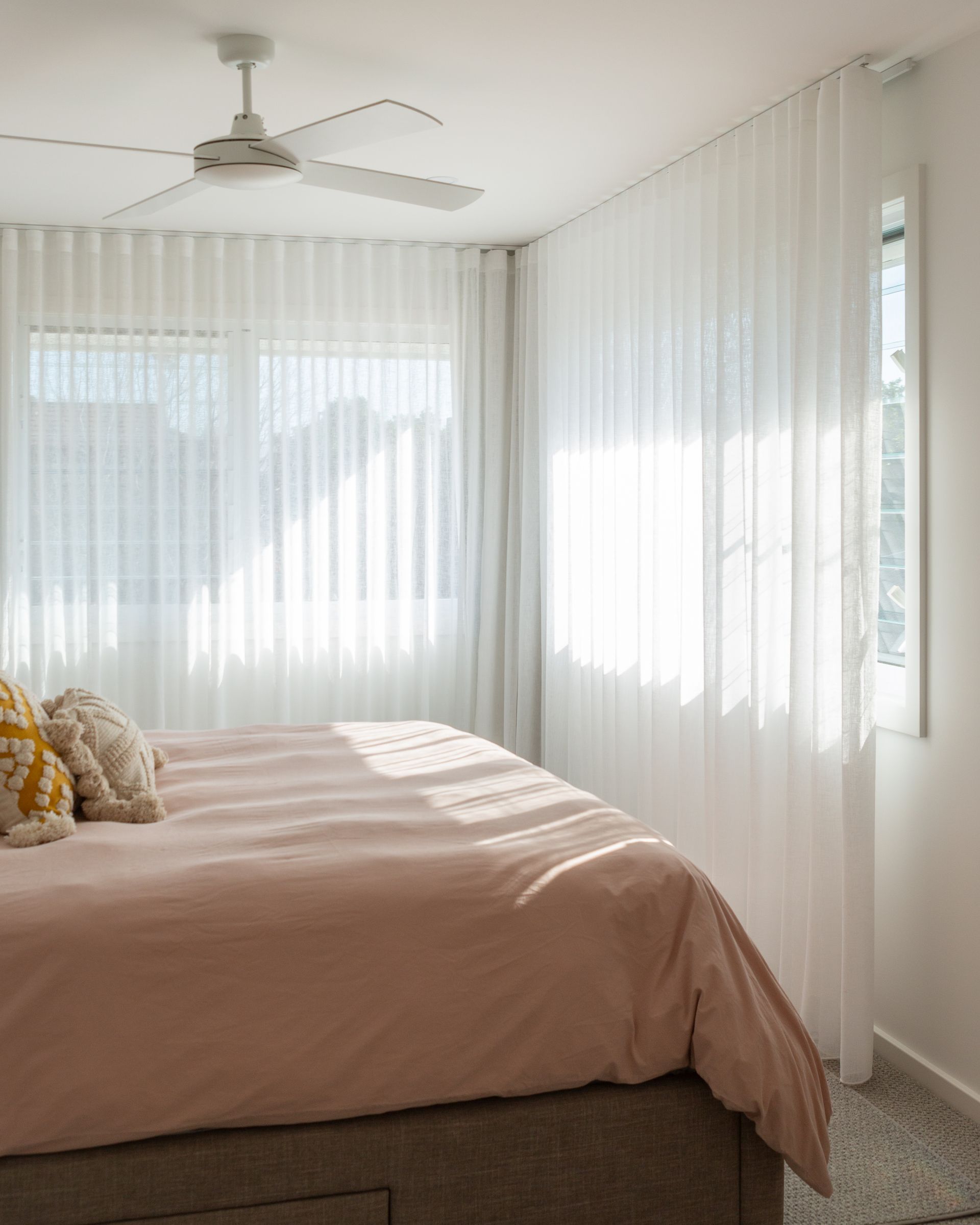 Bedroom with pink bed, sheer white curtains, and a ceiling fan. Sunlight streams through the windows.