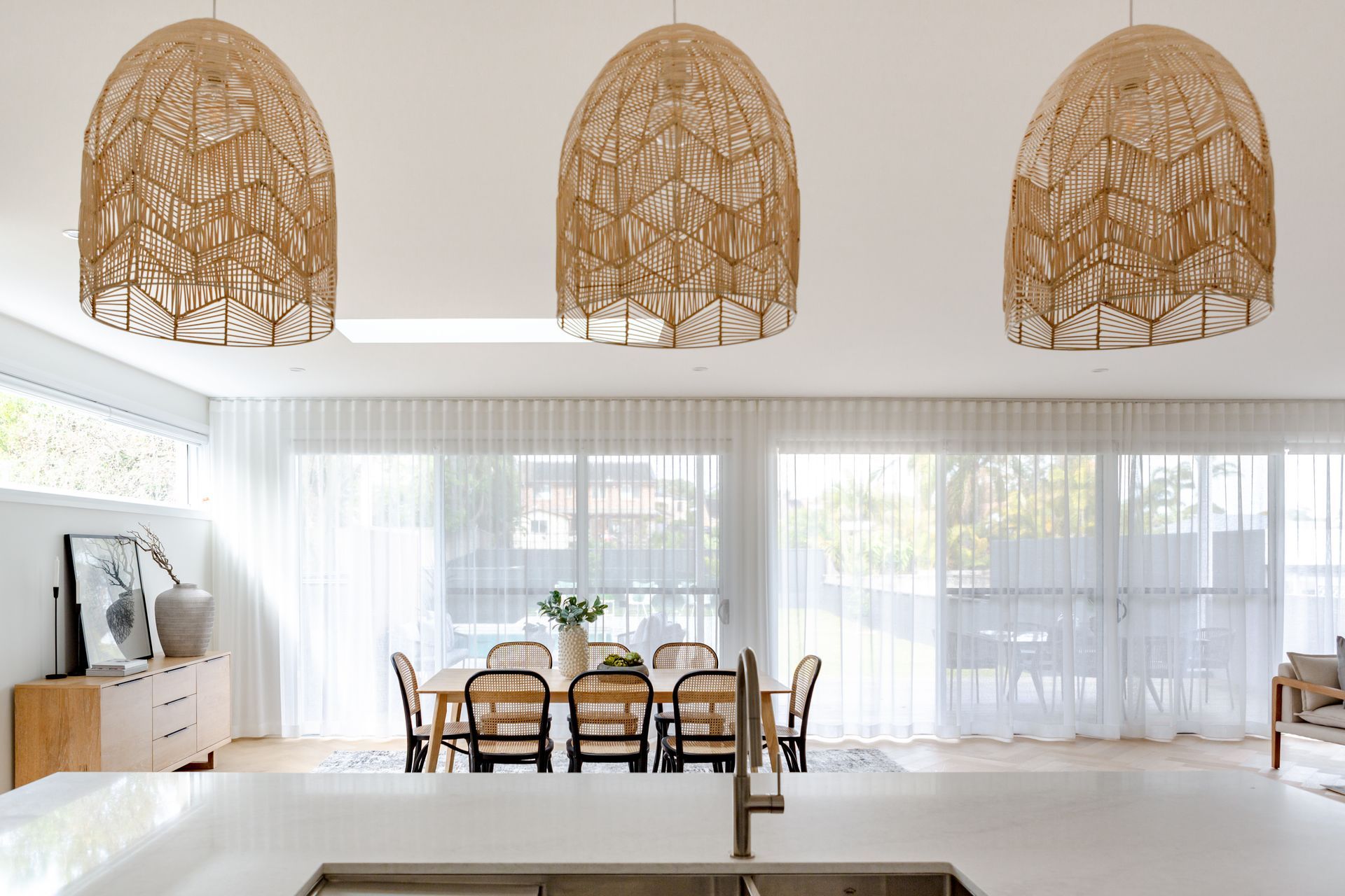 Dining room with woven pendant lights, table, chairs, sideboard, and sheer curtains.