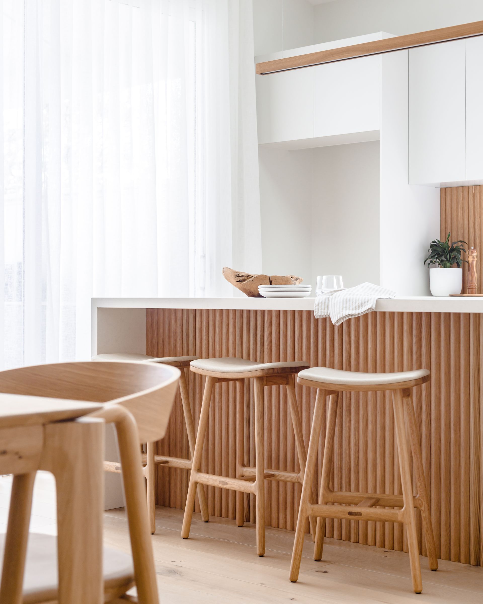 Wooden kitchen island with bar stools, light wood tones, white cabinets, and sheer curtains.