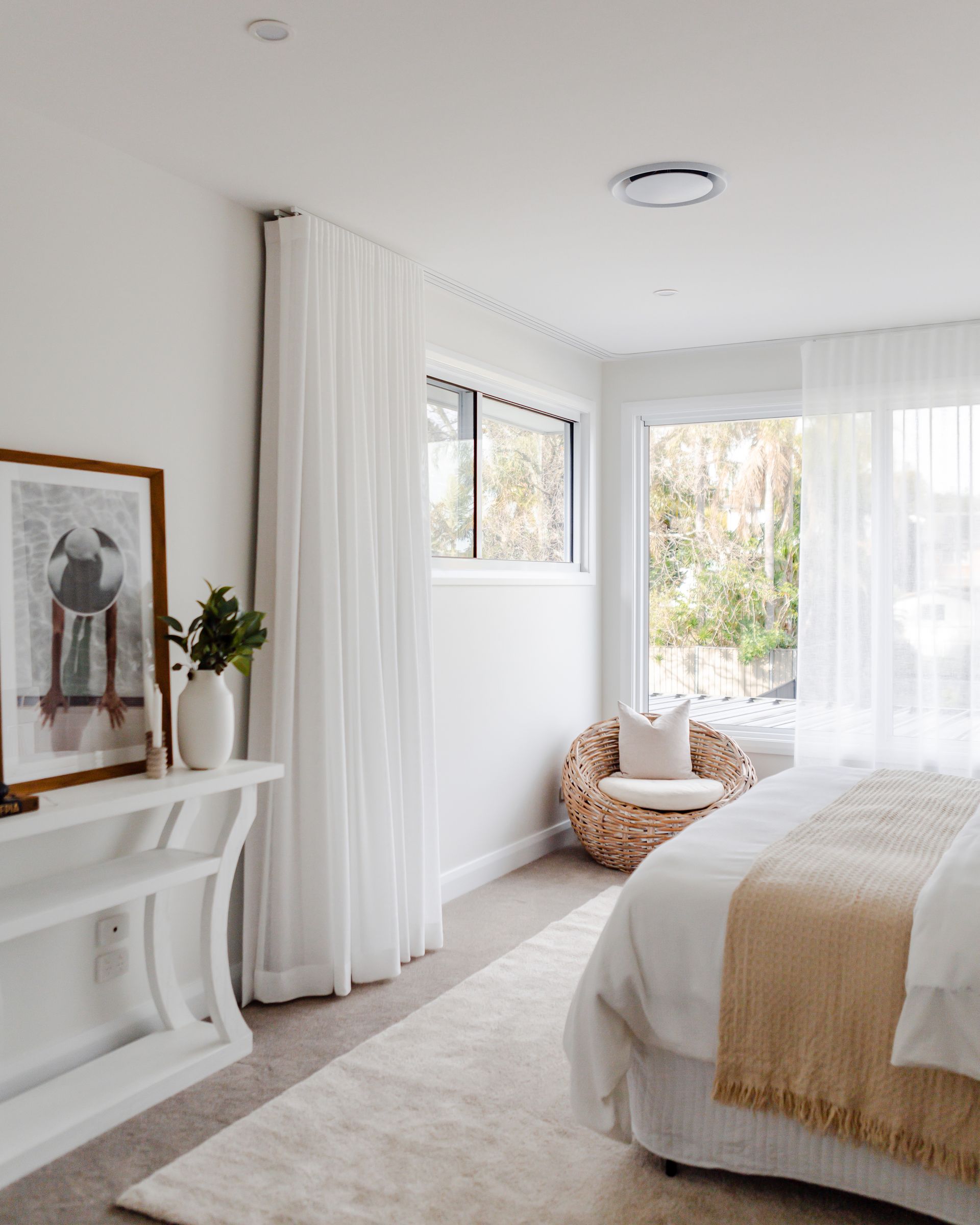 Cozy white bedroom with a bed, wicker chair, and a window overlooking greenery.