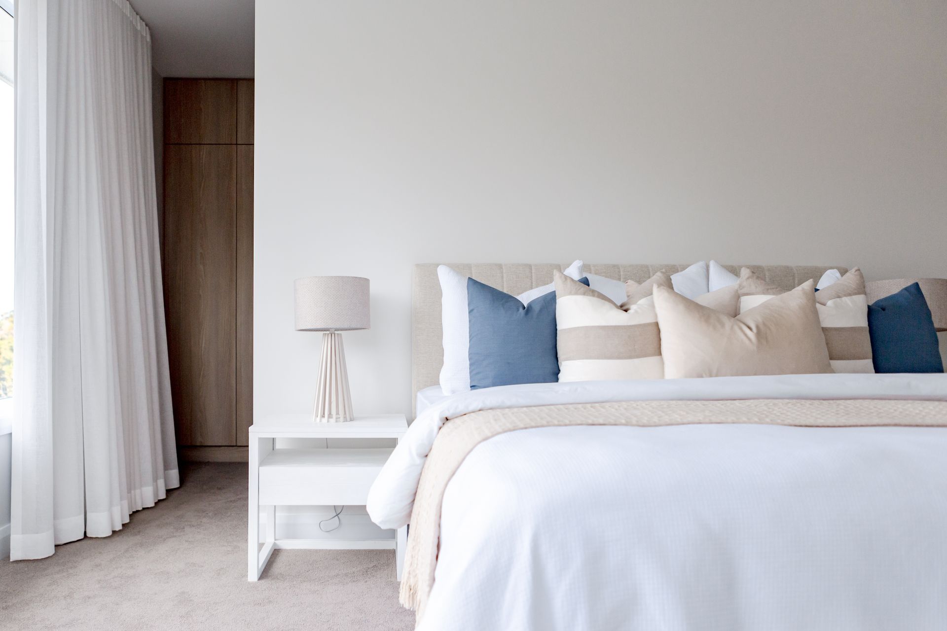 Bedroom with white bedding, blue and beige pillows, white nightstand, and sheer curtains.