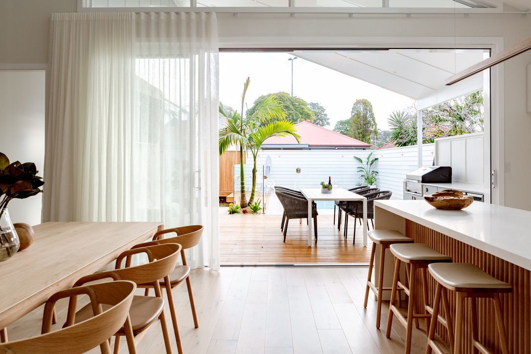 Dining room with open glass doors leading to a patio. Light wood, white walls, outdoor table, green plants.