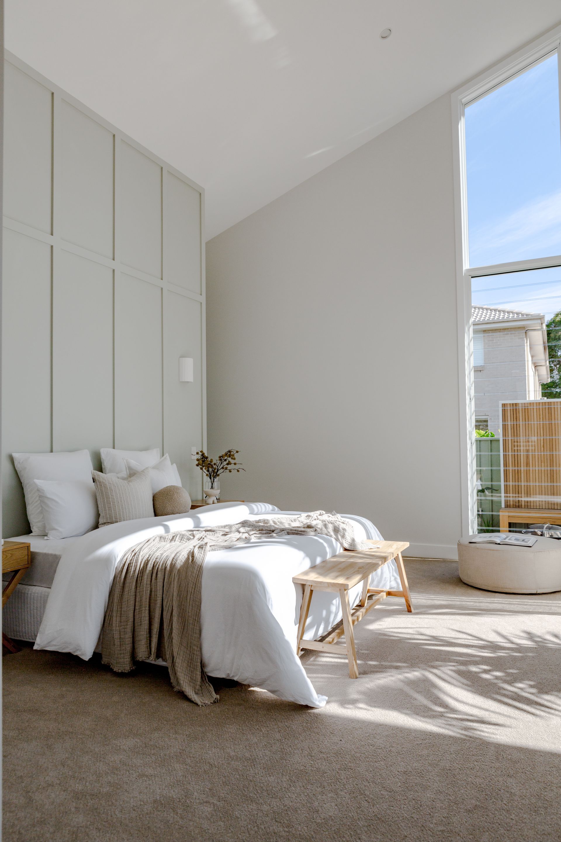 Bedroom with white bedding, neutral-colored wall panels, and large window casting shadows on the carpet.