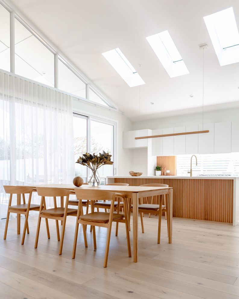 Light-filled dining area with wooden table, chairs, kitchen island, and skylights. Neutral tones with sheer curtains.