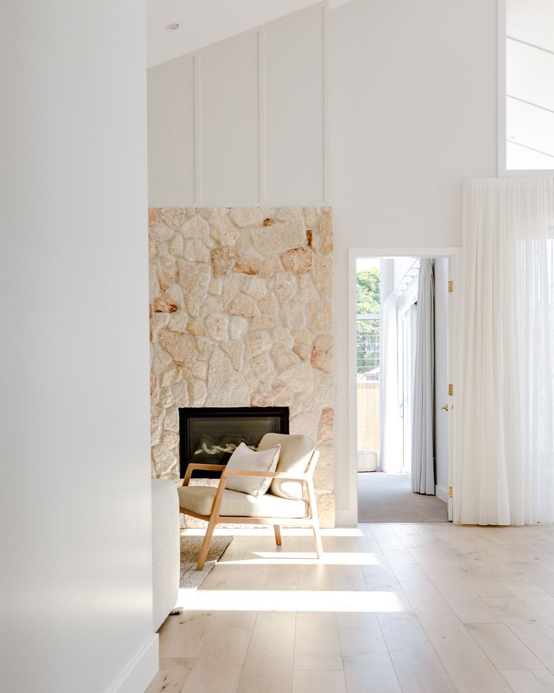 Living room with a stone fireplace, beige chair, and sheer white curtains; bright and airy.