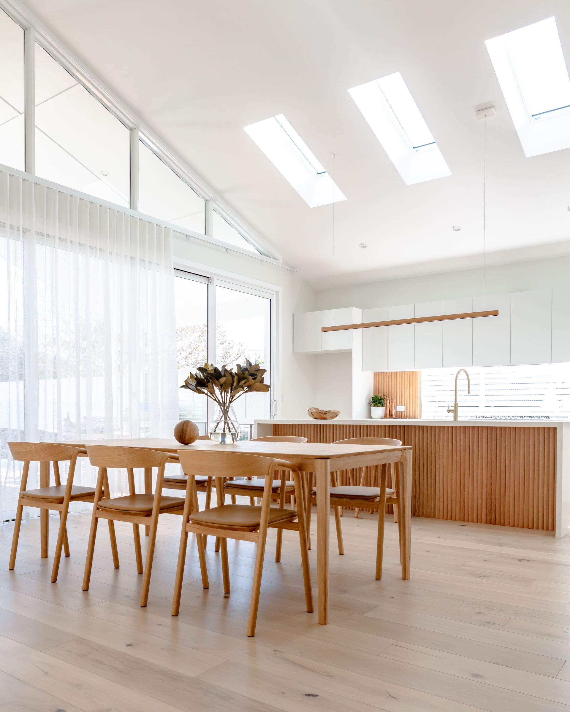 Light-filled dining room with a wooden table, chairs, and a kitchen featuring light wood cabinets.