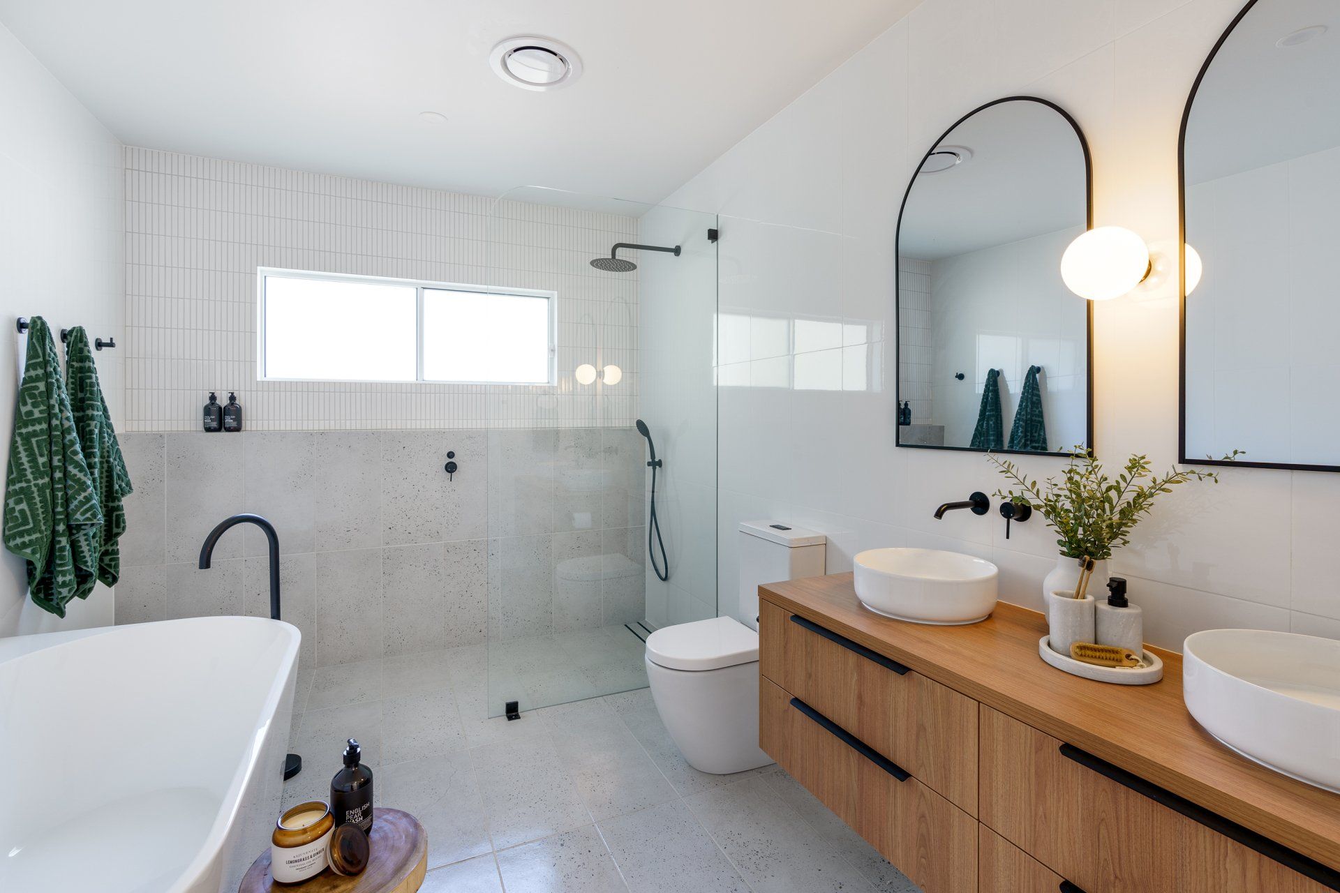 Modern bathroom with white walls, wood vanity, arched mirrors, and a freestanding tub.