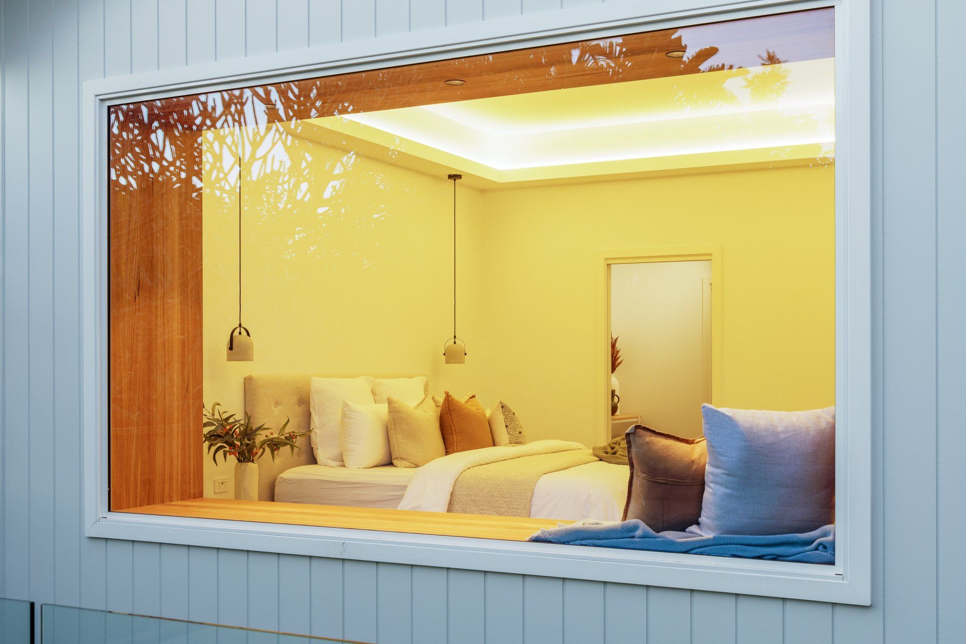 Bedroom interior seen through a window, featuring a bed with pillows, warm lighting, and wood paneling.