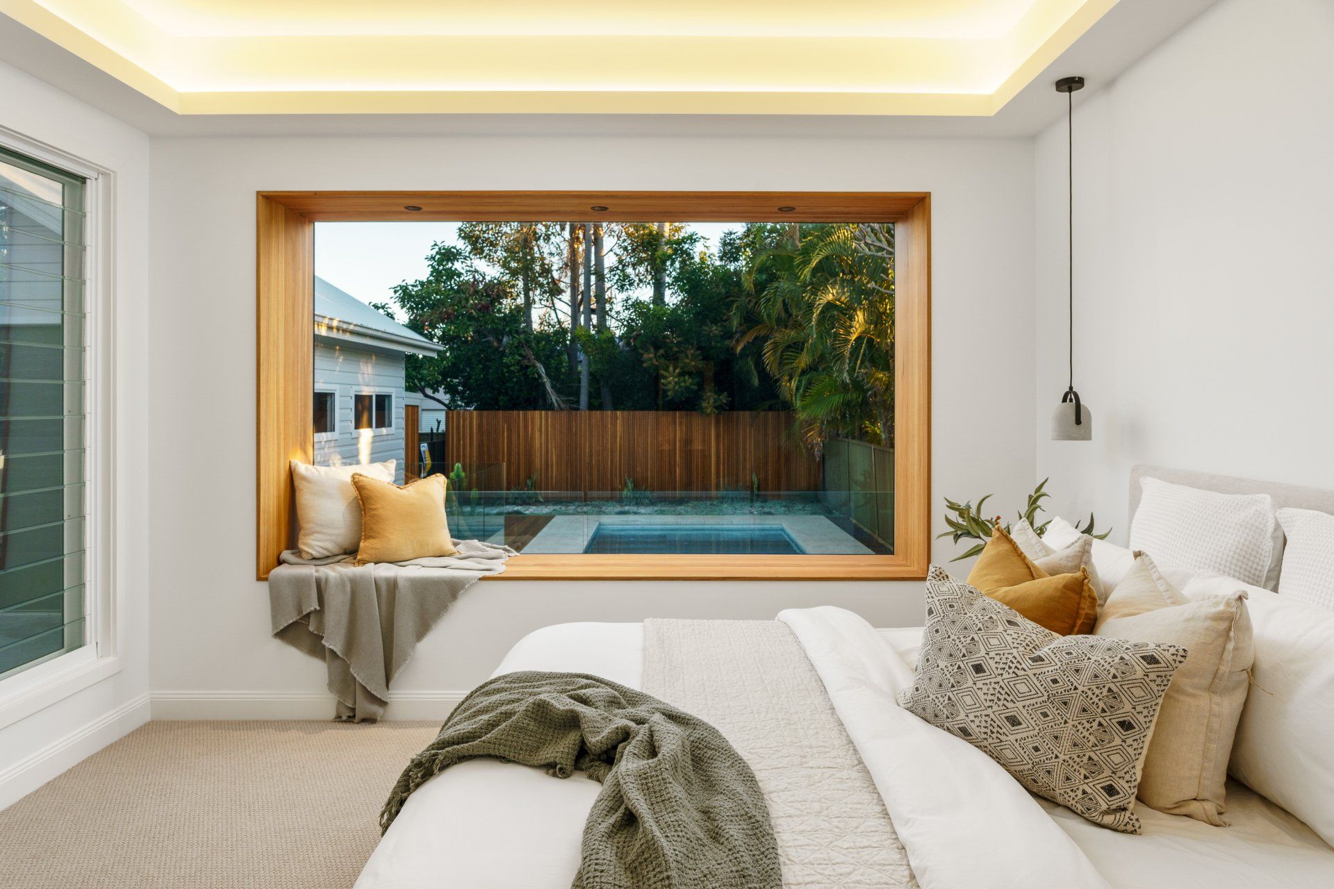 Bedroom with large window overlooking a pool and trees. Bed with pillows, window seat with pillows, and a blanket.