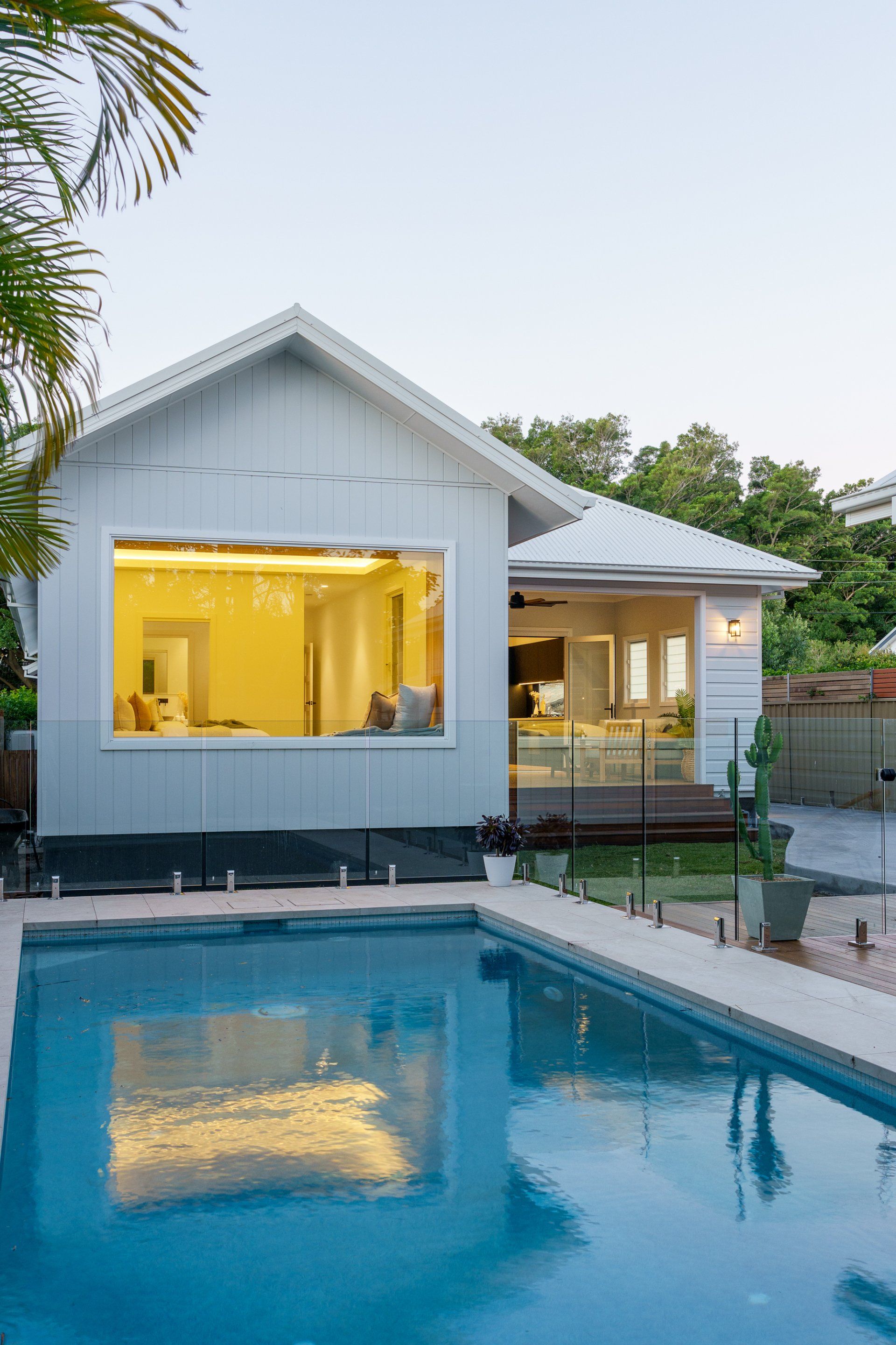 White house with pool, large window, yellow interior light, blue water.
