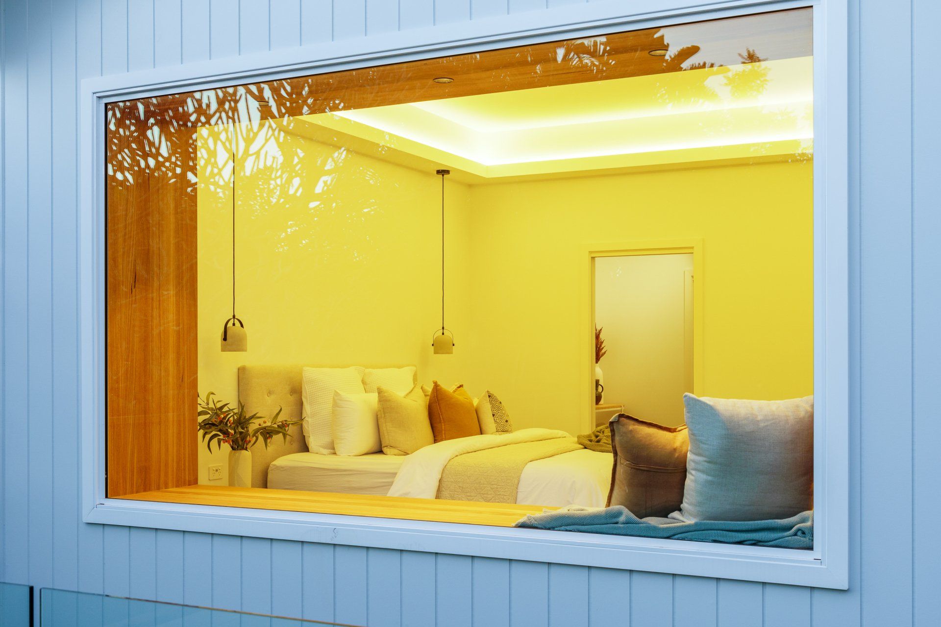 Bedroom interior seen through a large window, warm yellow lighting, a bed, pillows, and a window seat.