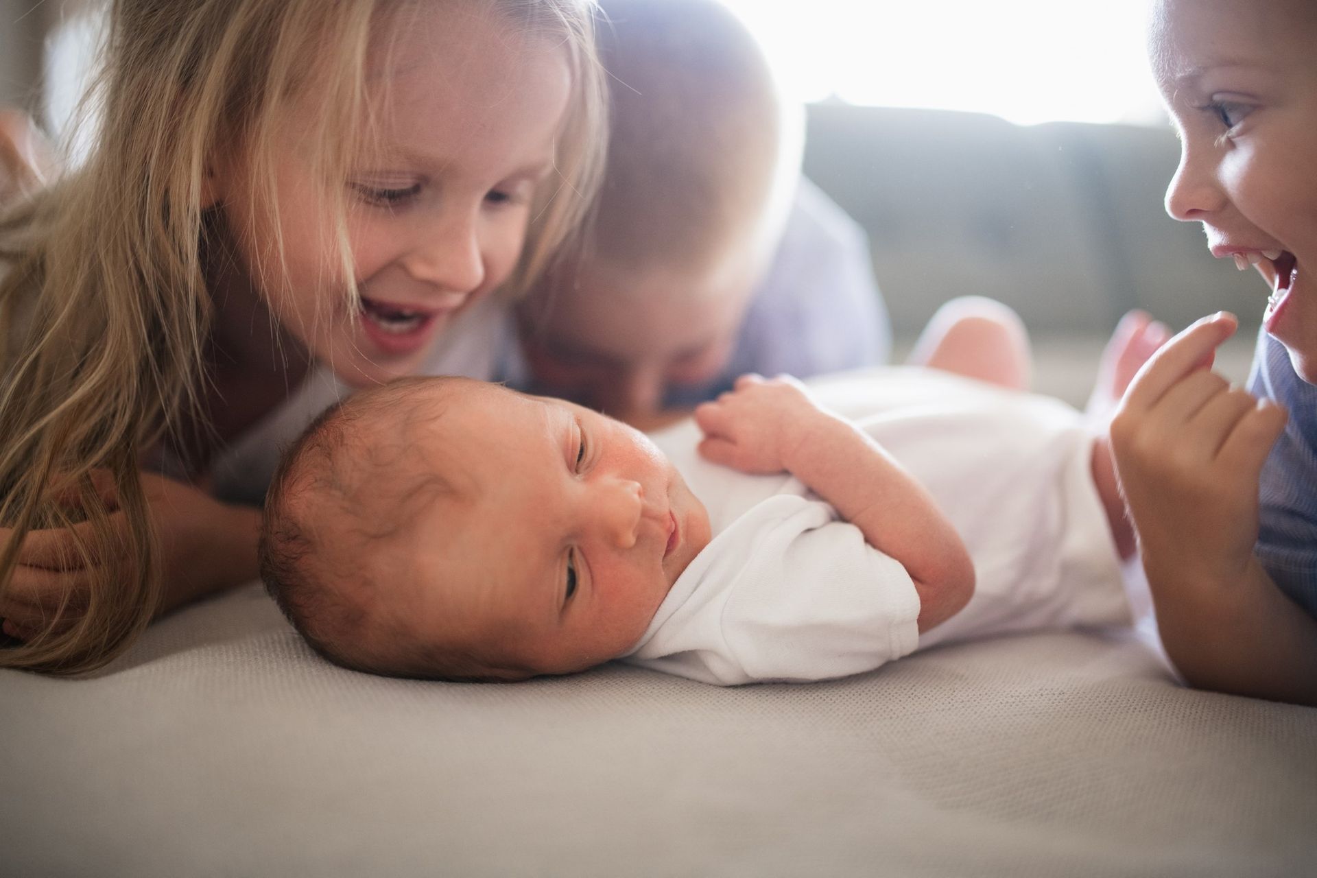 A little girl is laying on a sofa on her stomach and is looking at a small baby.