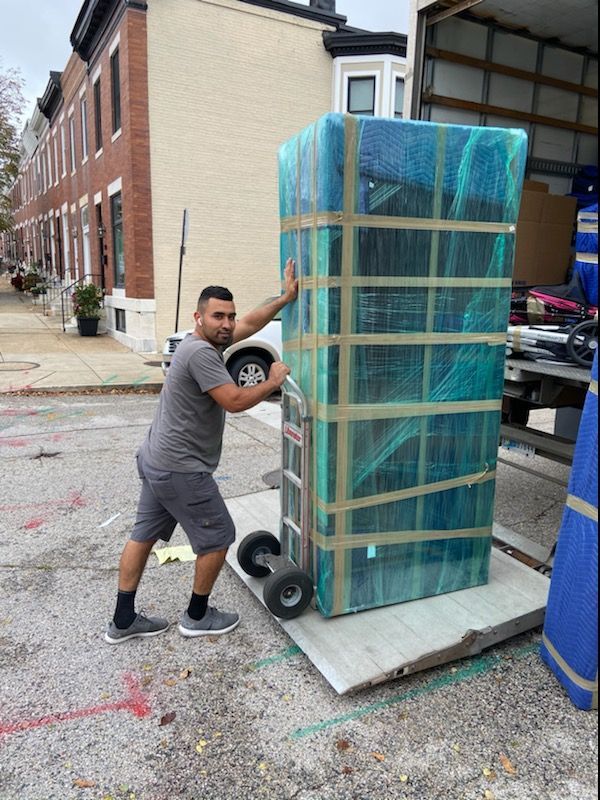 Man pushing a tall, wrapped mattress on a wheeled dolly beside parked cars on a city street