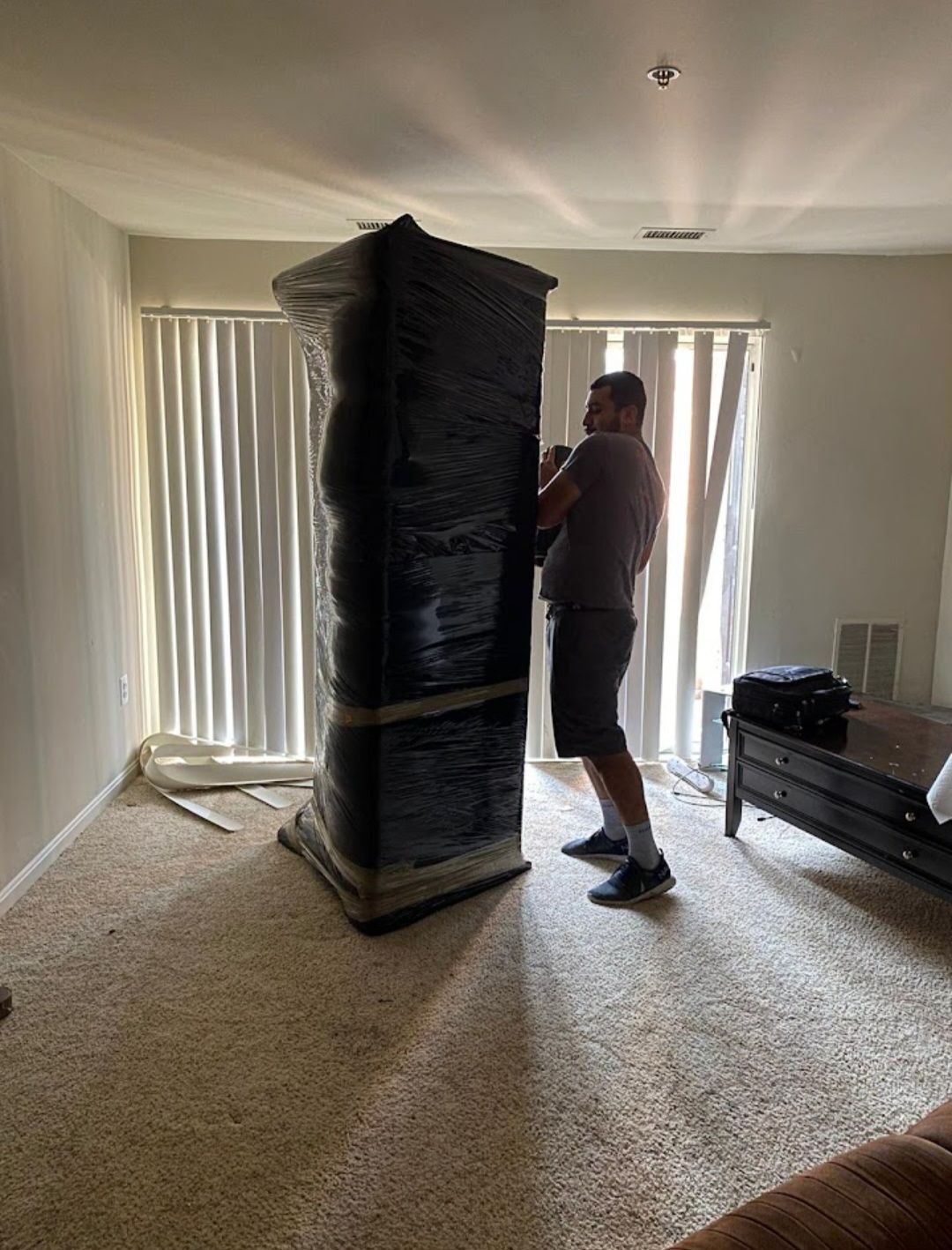 Man standing beside a tall black-wrapped cabinet in a carpeted living room with vertical blinds.