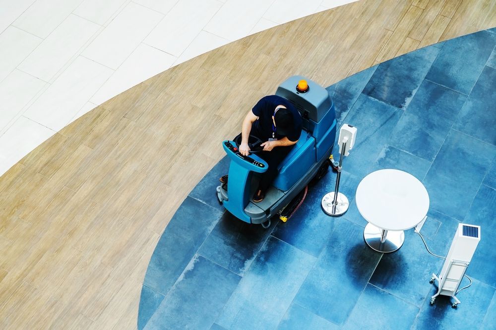 Person driving a floor cleaning machine on a blue-tiled floor next to a small table.