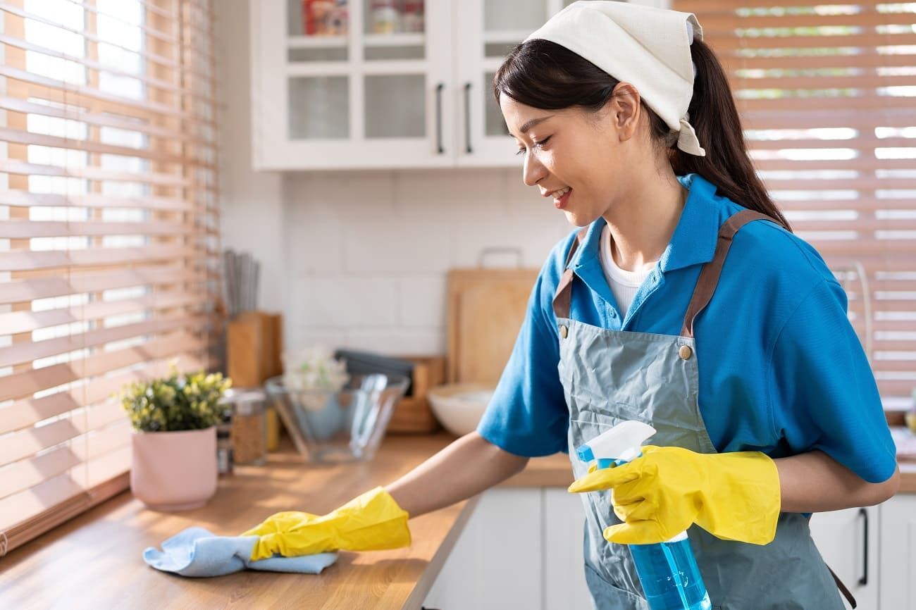 Woman wearing gloves, cleaning kitchen countertop with spray bottle and cloth.