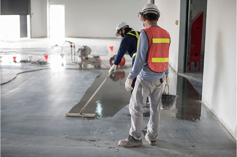 Two workers applying sealant to a concrete floor in a construction setting. One uses a squeegee.