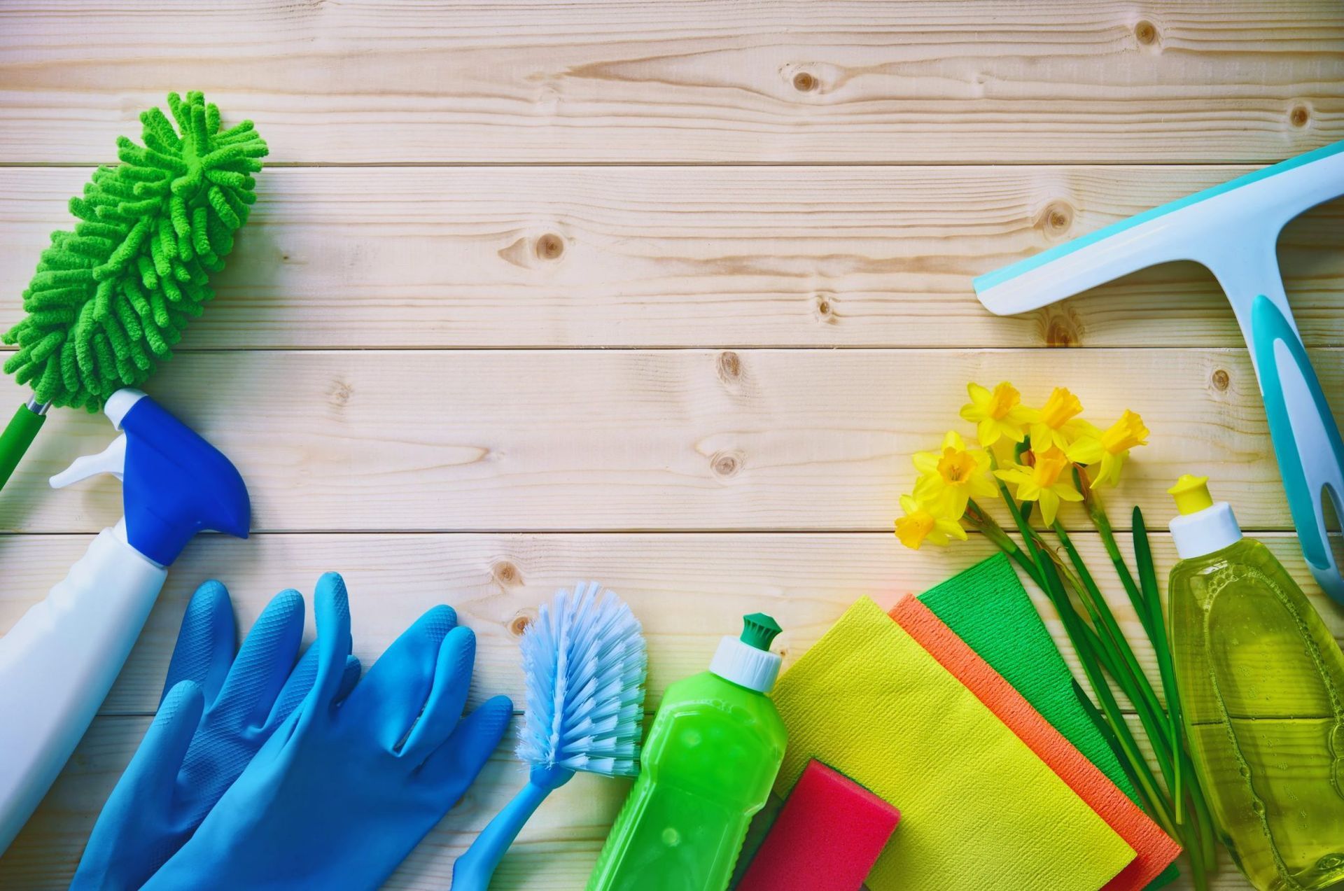 Cleaning supplies on a wooden surface: duster, spray bottle, gloves, brush, soap, cloths, squeegee, and daffodils.