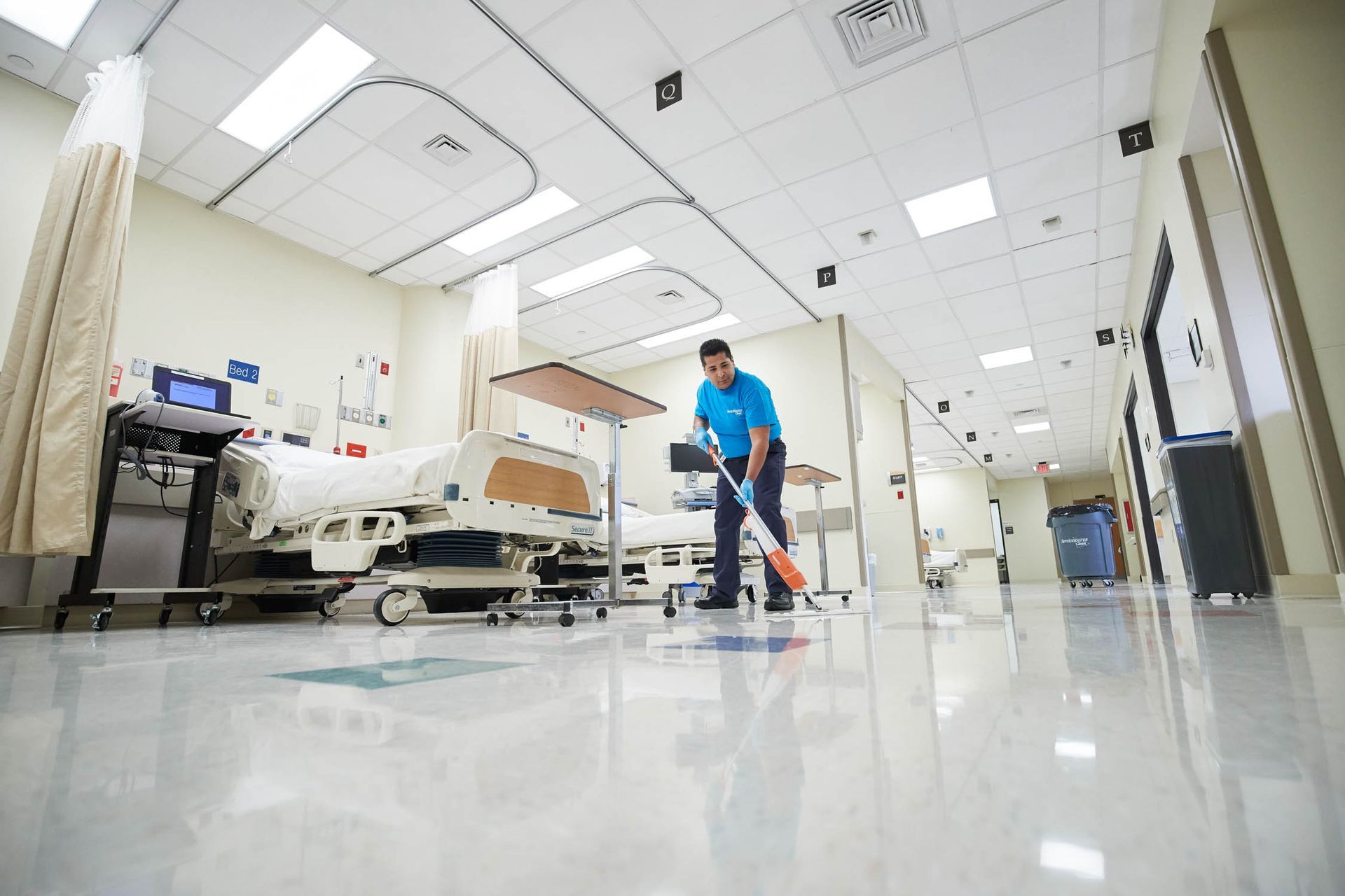Custodian mopping a shiny, white-floored hospital hallway with patient beds.
