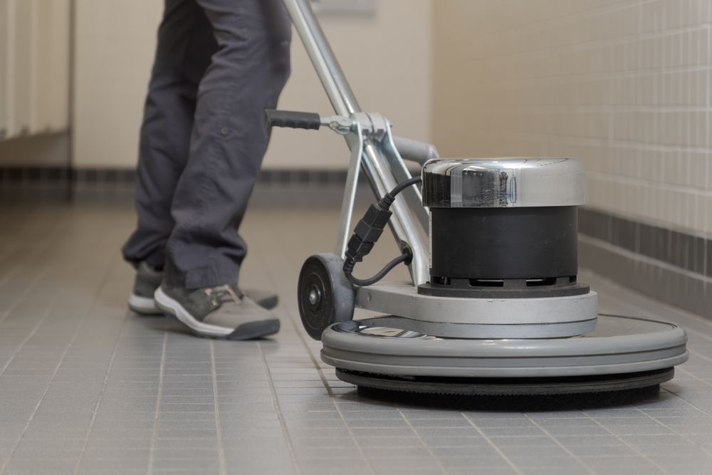 Person using a floor buffer to clean a tiled floor in a room with white walls.