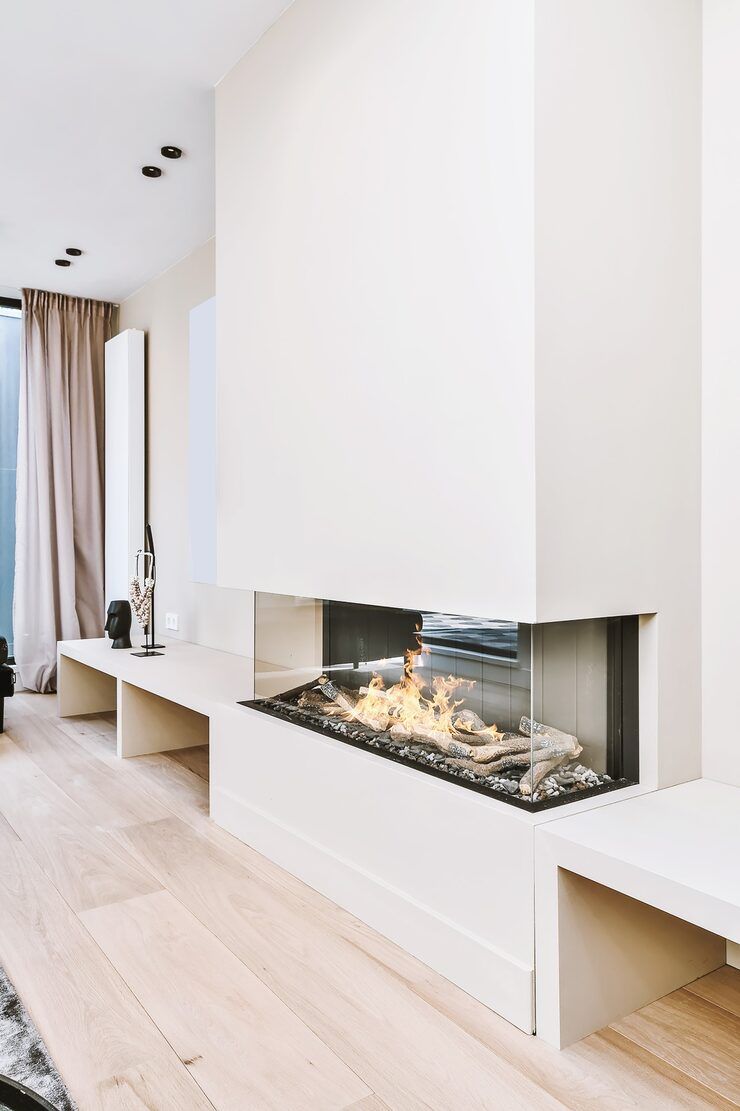 Modern white corner fireplace with glass front and burning logs.