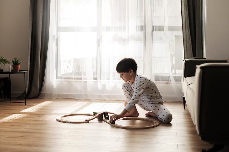 A child in pajamas playing with a wooden train set on a hardwood floor, by a sunlit window.