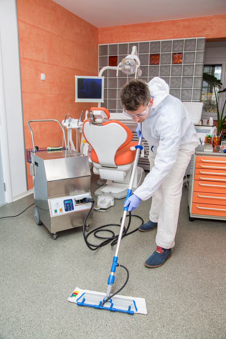Person in protective gear mops a dental office floor. Orange and white dental chair in the background.