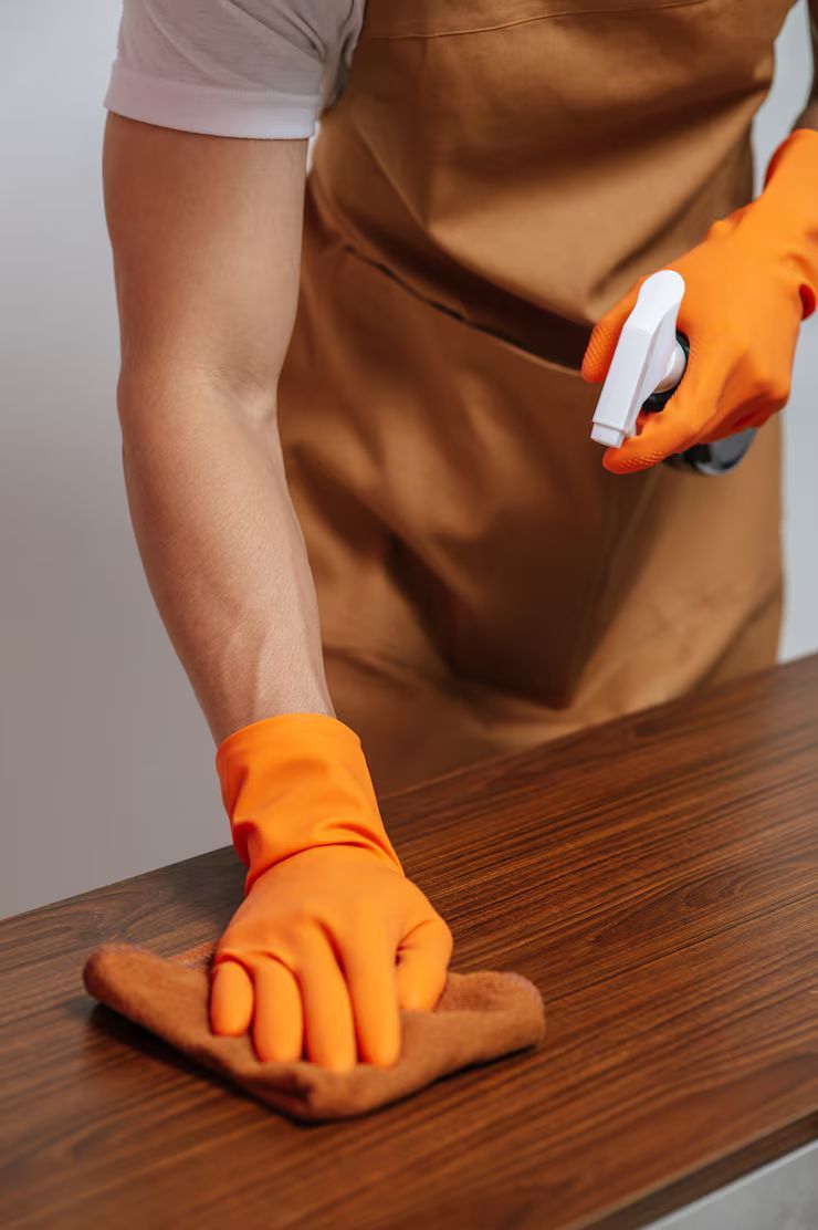 Person wearing orange gloves and apron, cleaning a wooden surface with a cloth and spray bottle.