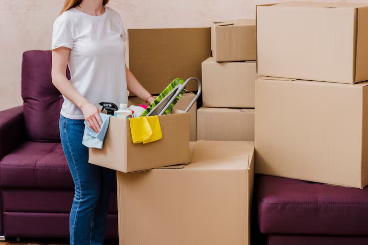 Woman in jeans and white shirt holding a box, surrounded by stacked moving boxes.
