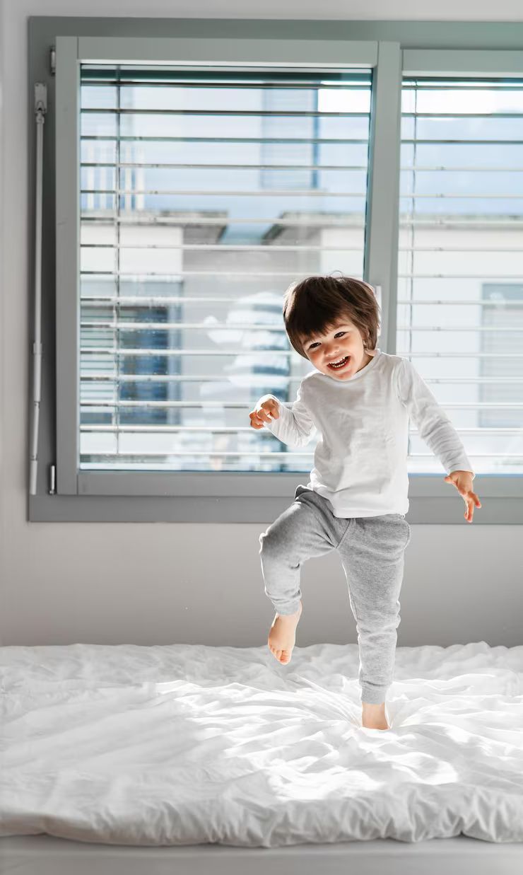 Happy child jumping on a white fluffy bed near a window.