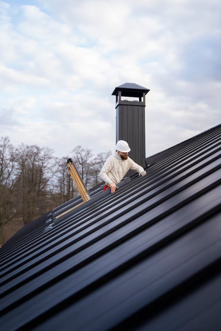 Person in white coveralls on a black metal roof next to a chimney, cloudy sky background.