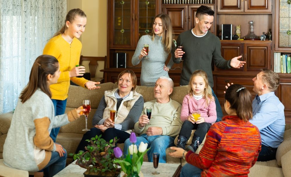 Family gathering indoors; people drinking and conversing on and around a sofa, with a bookcase in the background.