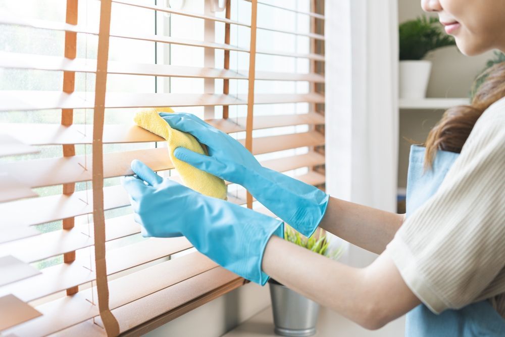 Woman in blue gloves cleaning wooden blinds with a yellow cloth.