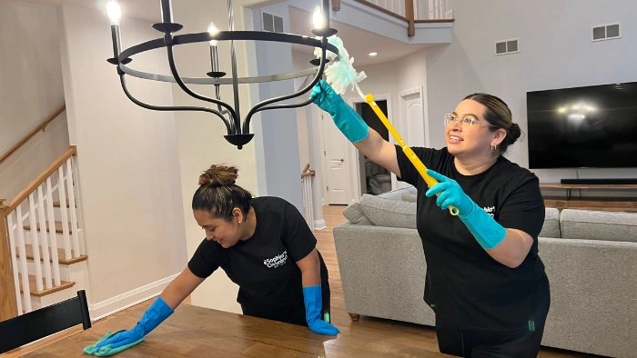 Two people cleaning a dining room. One dusts a chandelier, the other wipes a table. Both wear gloves.