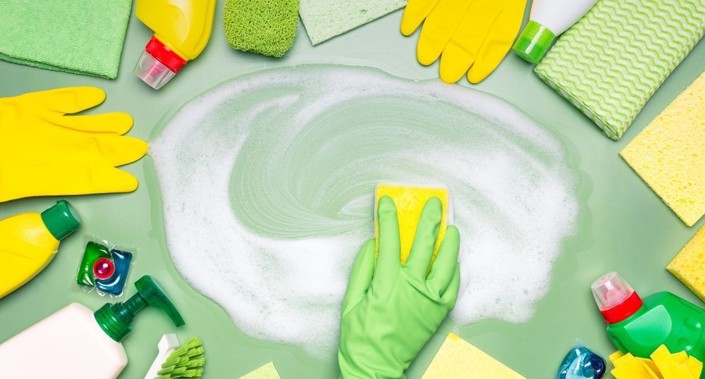 Green-gloved hand cleaning with yellow sponge, surrounded by cleaning supplies on a green surface.