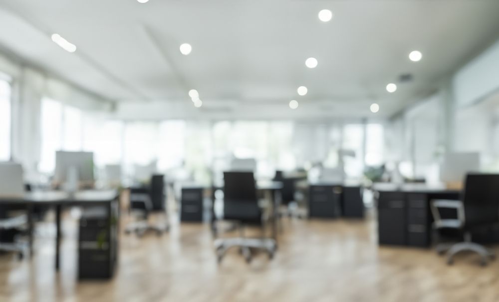 Blurred open office with desks, chairs, computers, and overhead lights.