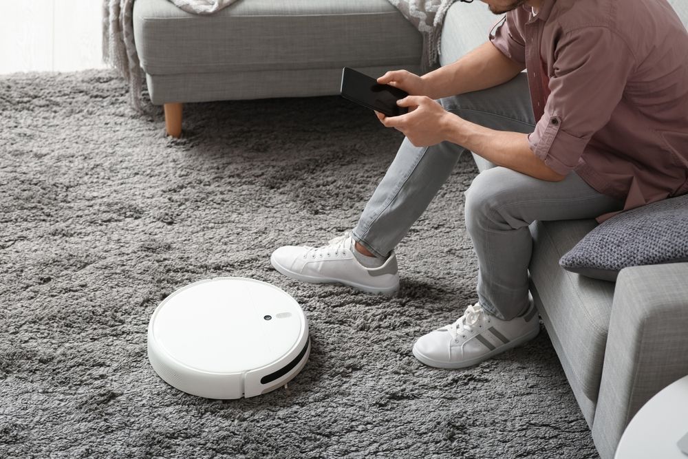 Person using a tablet controls a robotic vacuum on a gray carpet next to a sofa.