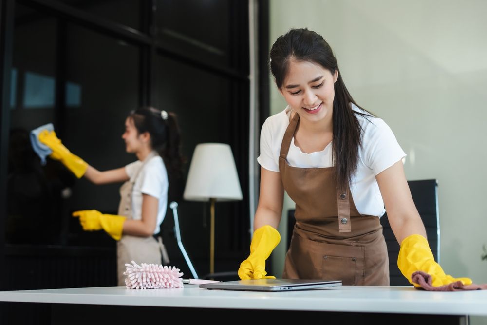 Two people wearing yellow gloves clean a room, one wiping windows, the other cleaning a desk.