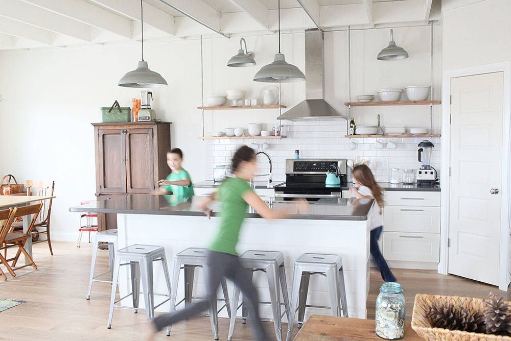 Children running around a kitchen island. White cabinets, stainless steel range hood, wooden shelves, and pendant lights.