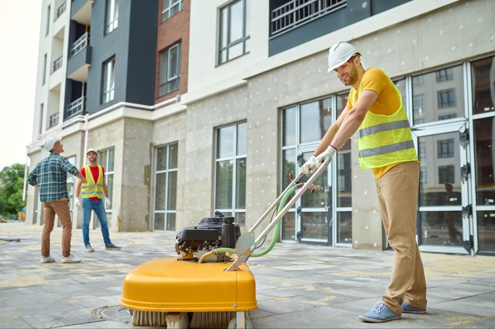 Construction worker using a power brush on a walkway, other workers watch. Building in background.