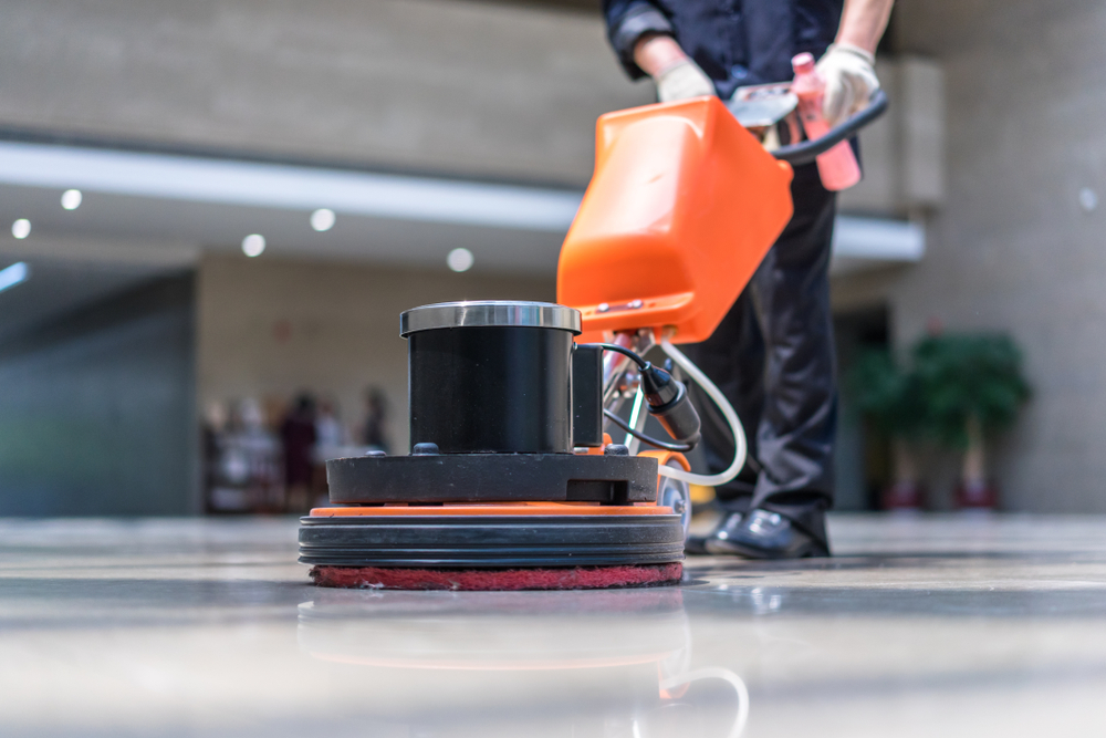 Person using an orange floor buffer to clean a polished floor in a bright lobby.