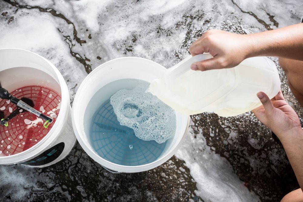 Person pouring liquid from a jug into a bucket filled with soapy water, another bucket nearby.