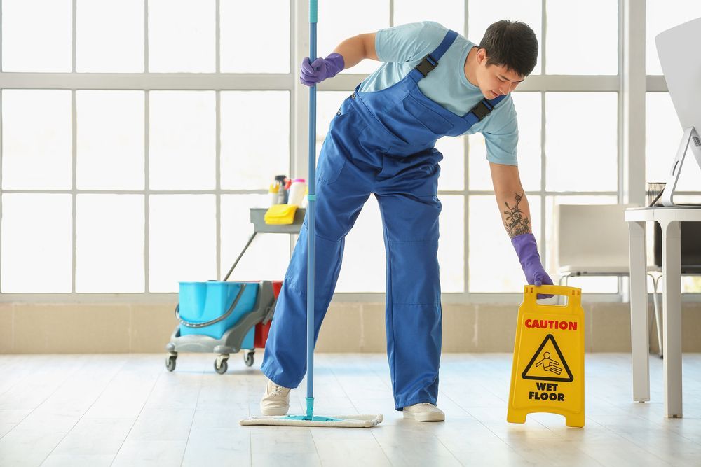 Man in blue overalls mops a floor, placing a “Wet Floor” sign near a desk.
