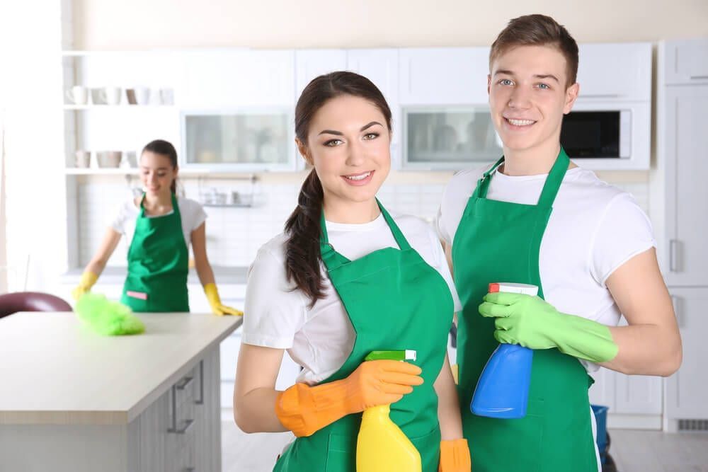 Three people wearing green aprons and gloves cleaning a white kitchen. Two smile at the camera, holding spray bottles.